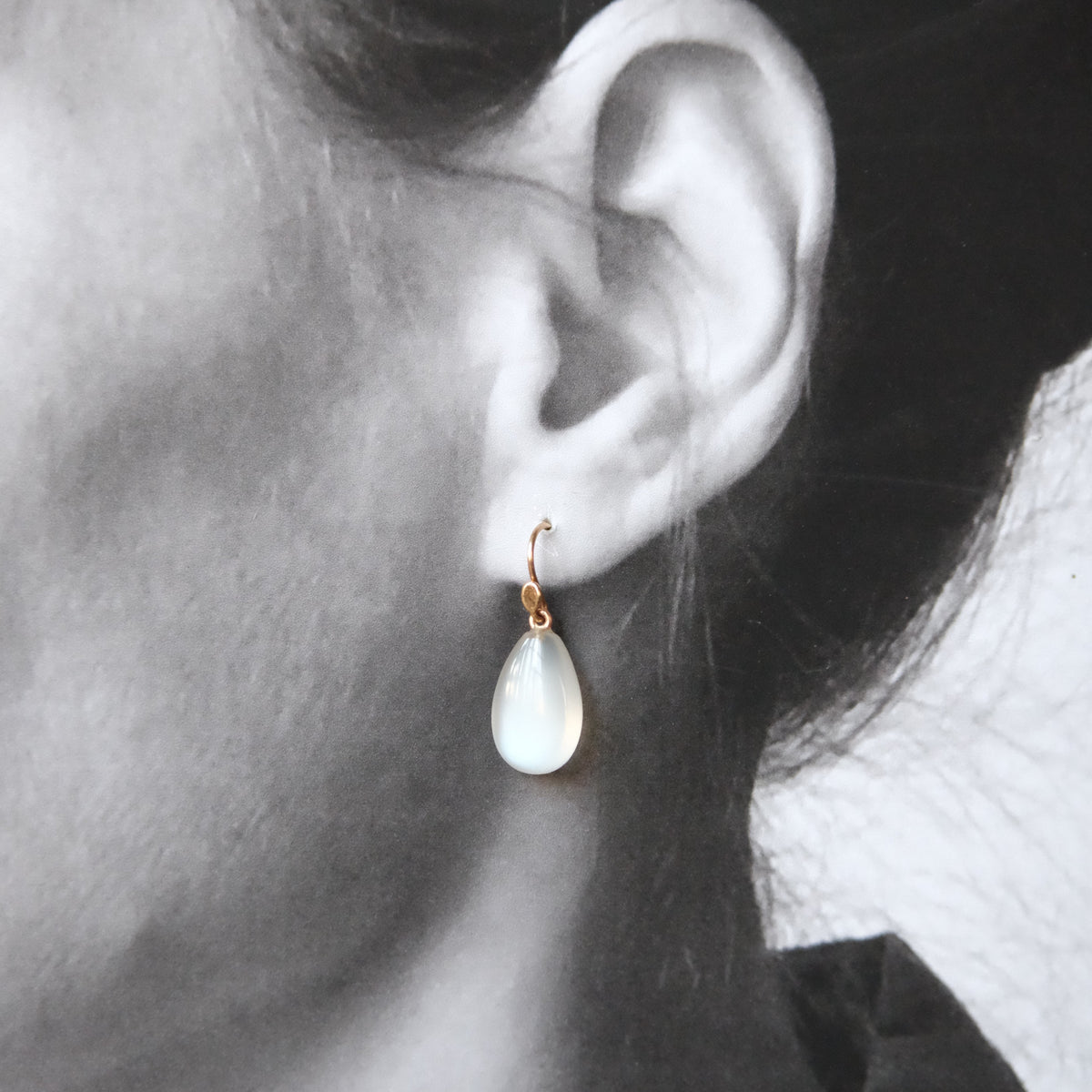 A close-up of a person's ear wearing a small gold hoop earring with a teardrop-shaped milky white stone drop.