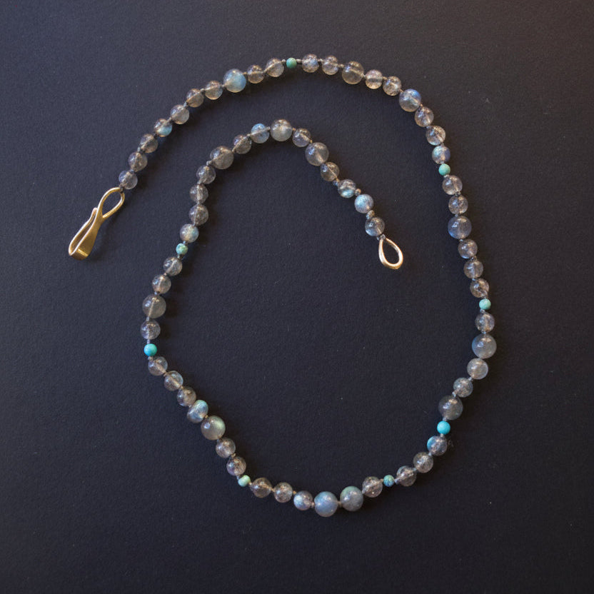 A beaded necklace with faceted gray beads that shimmer with iridescent blue and green flashes, accented by small turquoise-blue beads and gold-toned metal hardware, displayed on a dark background.