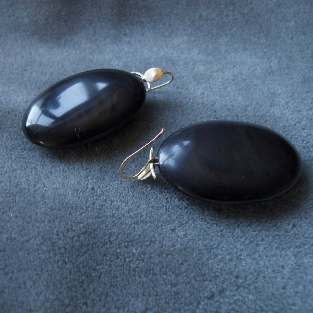 A pair of oval black stone drop earrings with gold fishhook wires and small pearl-like accent beads, displayed on a textured dark gray fabric background.