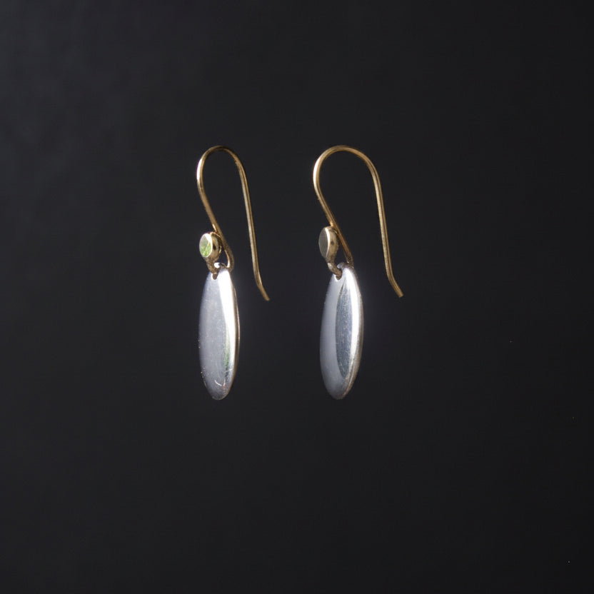 A pair of gold-tone drop earrings with oval silver-toned pendants, hanging from a thin wire against a dark black background.