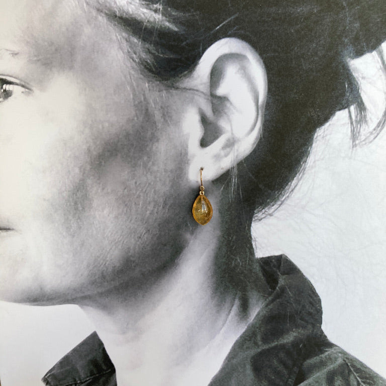 A close-up side profile of a woman wearing a small gold teardrop dangle earring, shown against a black-and-white portrait background.