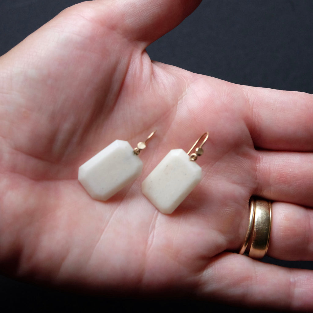 A pair of small rectangular cream-colored stone drop earrings with gold hooks, resting on an open hand against a dark background.
