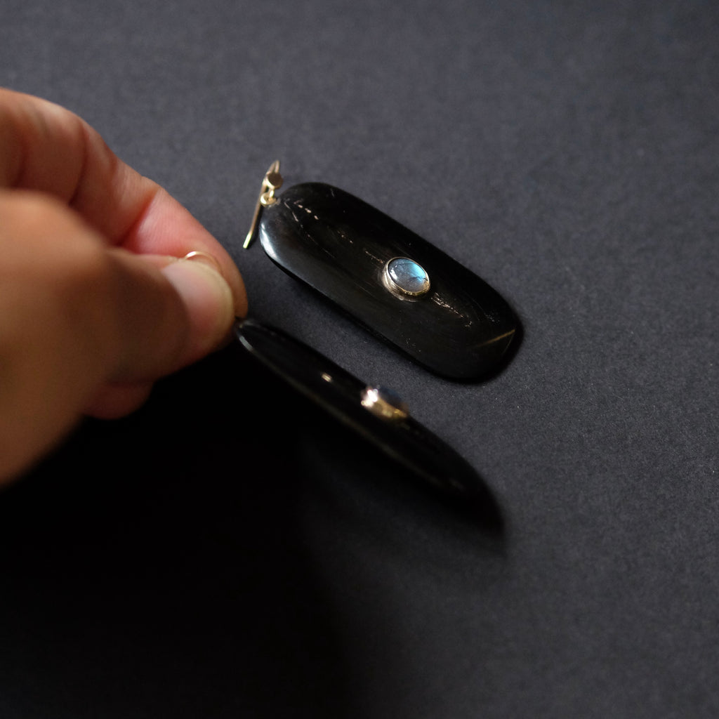 A hand holds a small black oval case or pendant with a gold-tone clasp and a round iridescent stone, next to another matching piece on a dark gray surface.