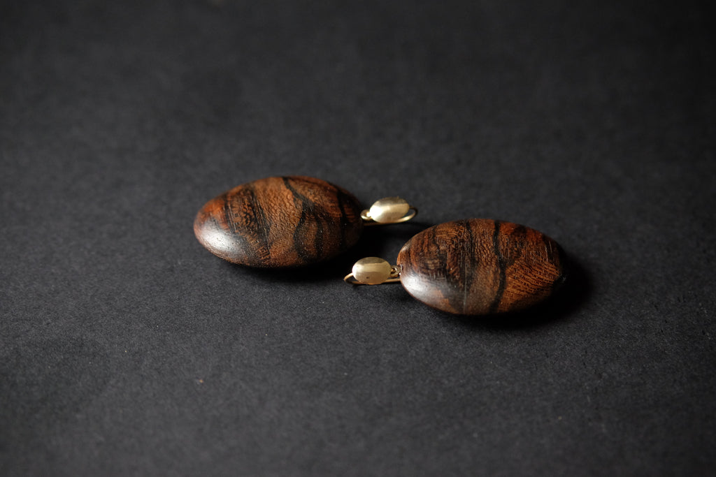 A pair of handmade drop earrings made from polished dark brown wood beads with natural grain patterns, connected by small gold-colored metal spacers, set against a dark textured background.