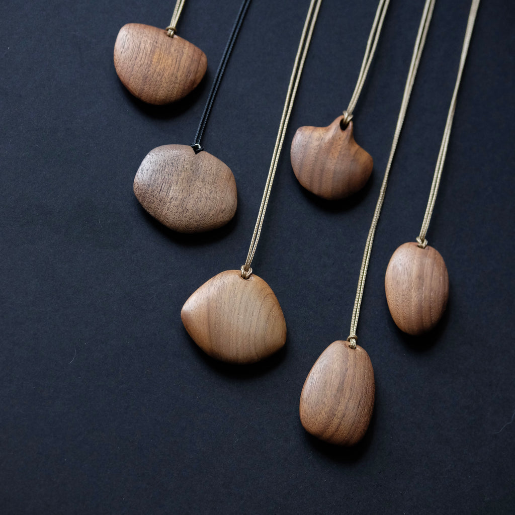 Five wooden pebble-shaped necklaces with different wood tones and cord colors, arranged diagonally on a dark textured background.