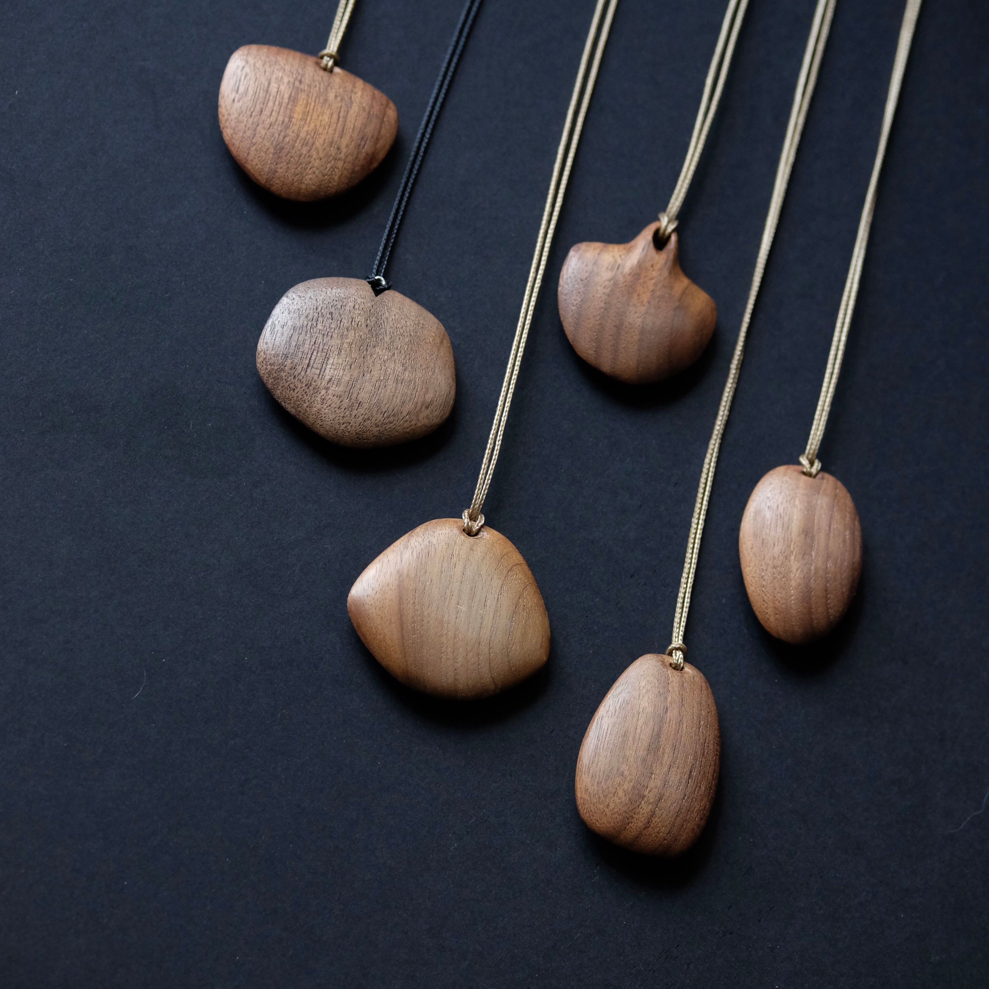 Five wooden pebble-shaped necklaces with different wood tones and cord colors, arranged diagonally on a dark textured background.