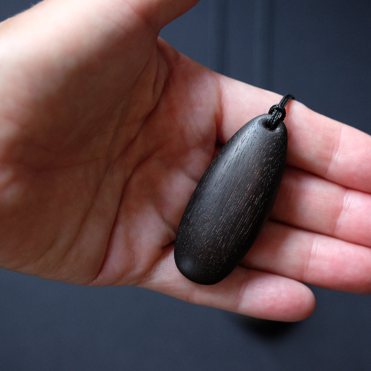 A dark, polished wooden pendant with a visible wood grain texture, held in the palm of a hand against a dark gray background.