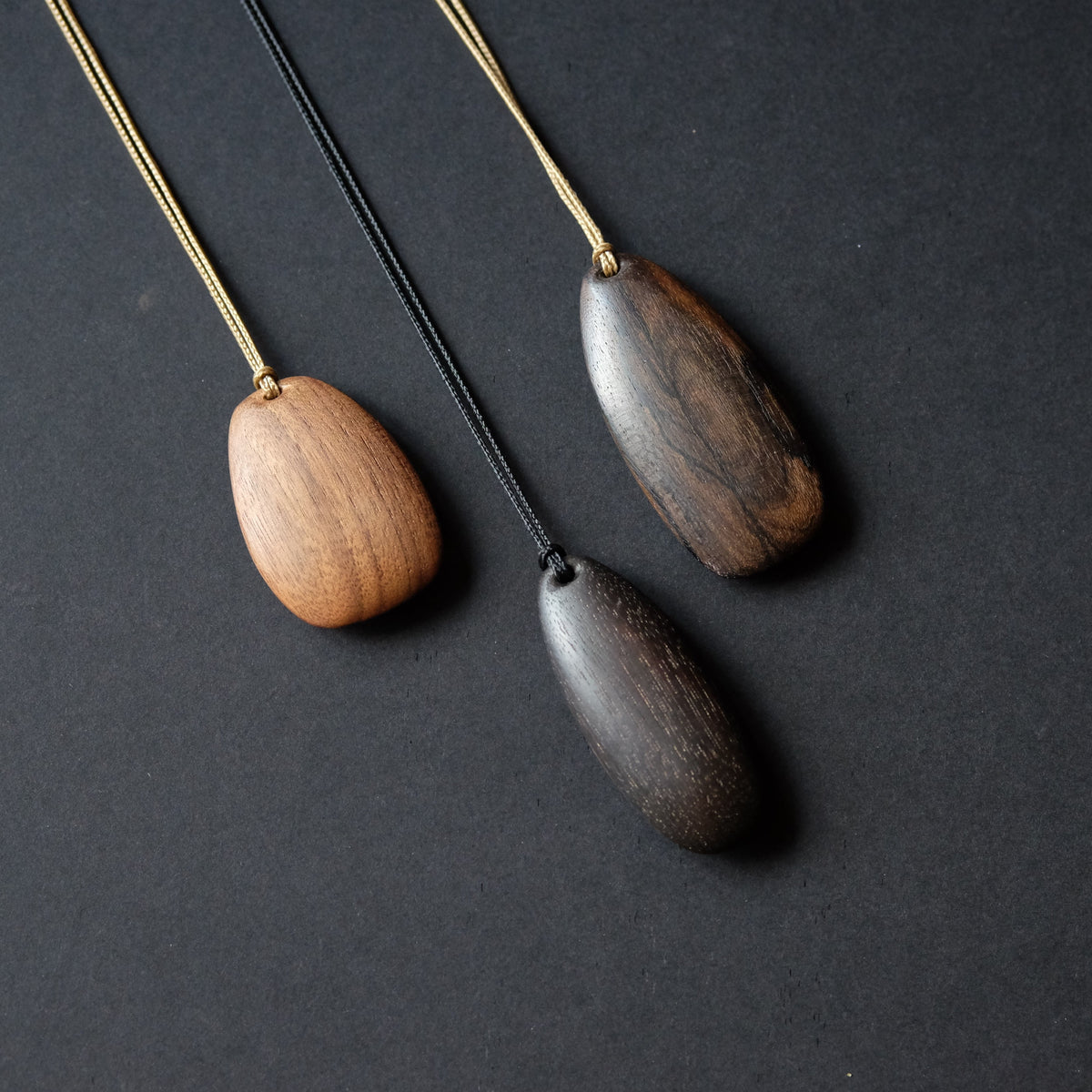 Three wooden pendant necklaces with different wood tones and black cords, arranged diagonally on a dark textured background.