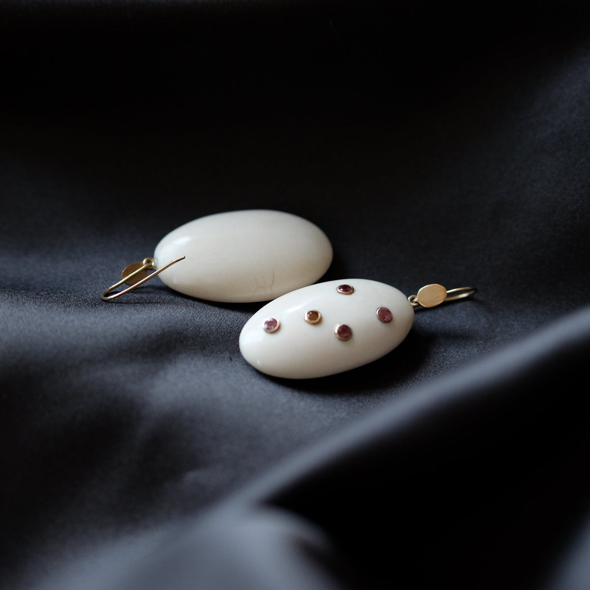 Two white oval pendant earrings with small gold hooks and tiny red gemstone accents, resting on draped black fabric.