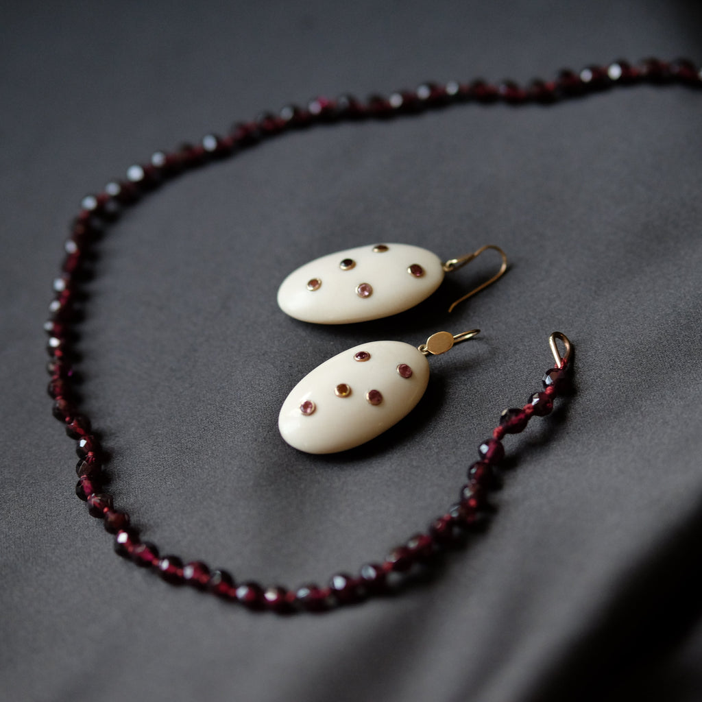 A jewelry set featuring a beaded necklace and matching drop earrings. The necklace and earrings are made of dark red faceted beads and cream-colored oval pendants decorated with small red dots, hanging from gold-toned hooks, all arranged on a dark gray fabric background.