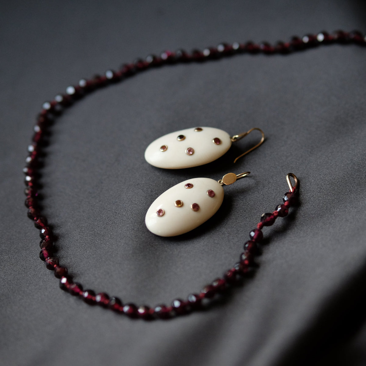 A jewelry set featuring a beaded necklace and matching drop earrings. The necklace and earrings are made of dark red faceted beads and cream-colored oval pendants decorated with small red dots, hanging from gold-toned hooks, all arranged on a dark gray fabric background.