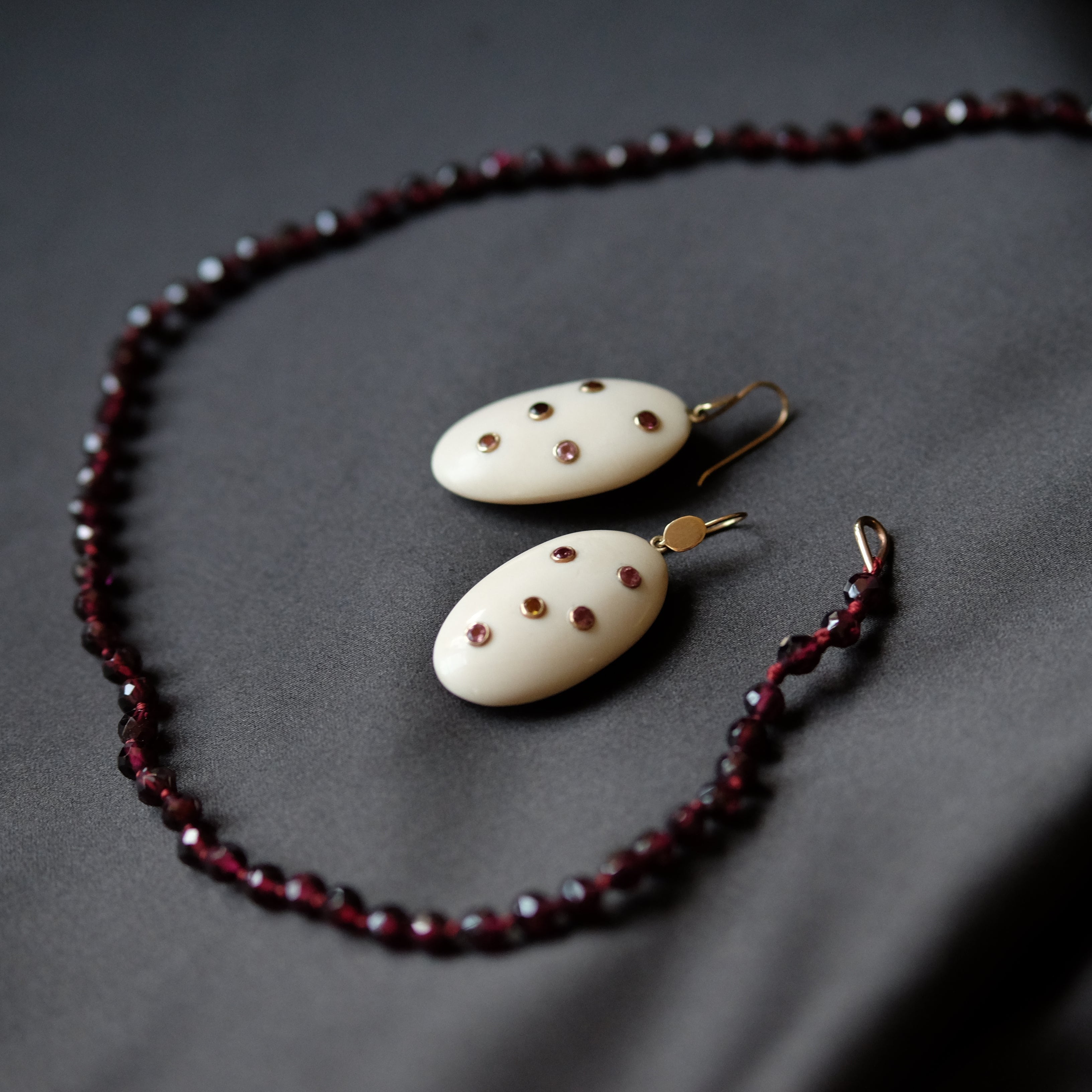 A jewelry set featuring a beaded necklace and matching drop earrings. The necklace and earrings are made of dark red faceted beads and cream-colored oval pendants decorated with small red dots, hanging from gold-toned hooks, all arranged on a dark gray fabric background.