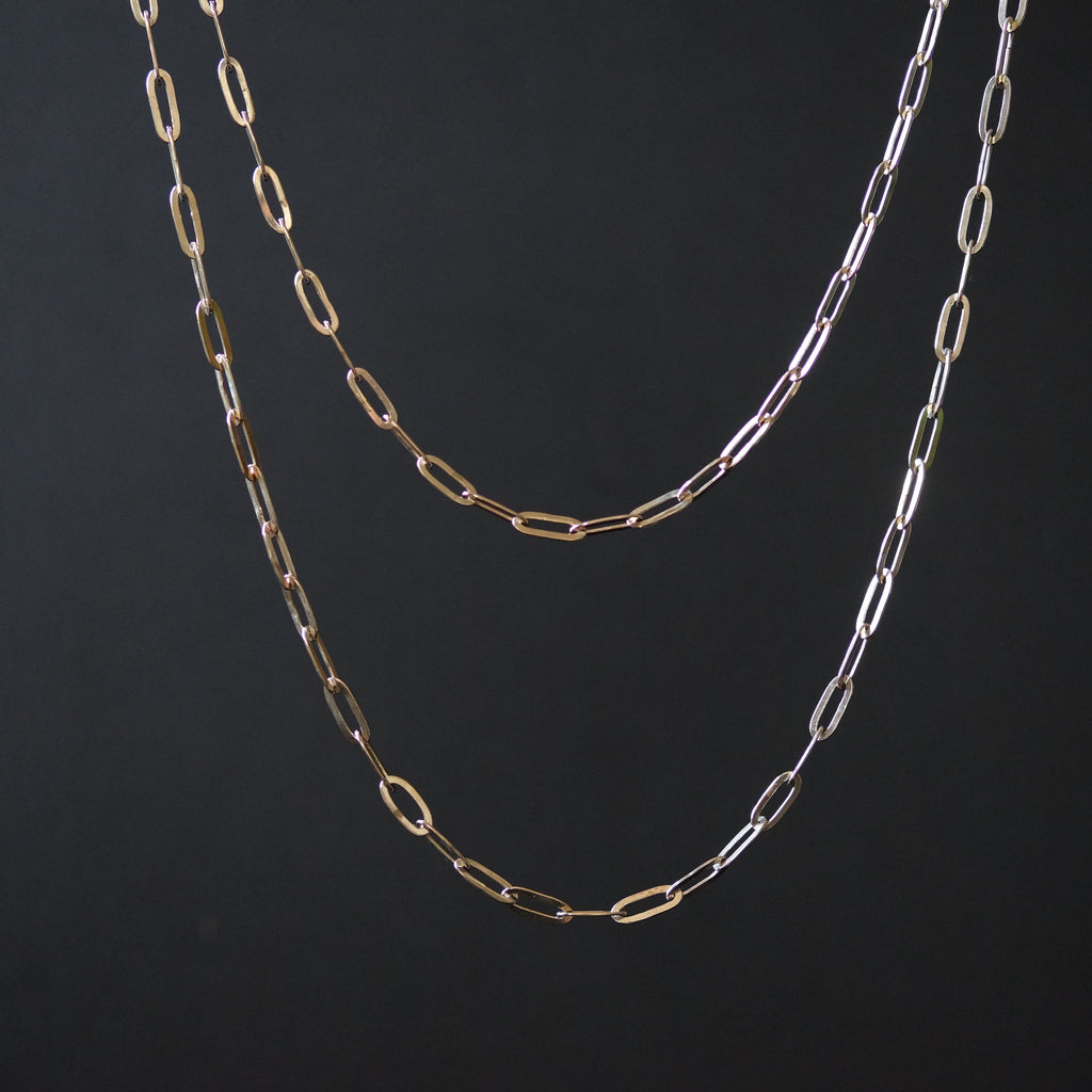 Two paperclip-link chain necklaces in gold and silver tones, hanging against a dark black background.