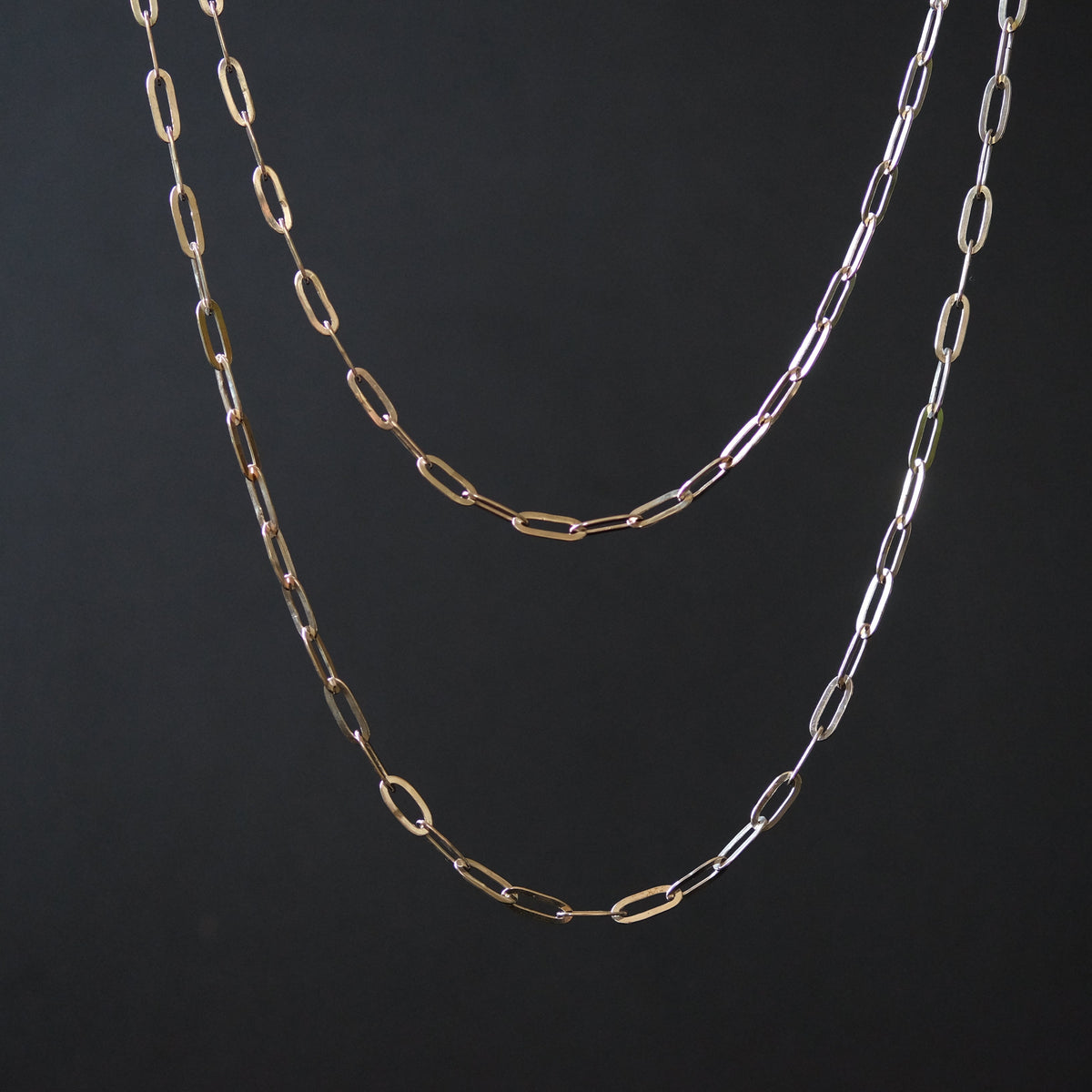 Two paperclip-link chain necklaces in gold and silver tones, hanging against a dark black background.