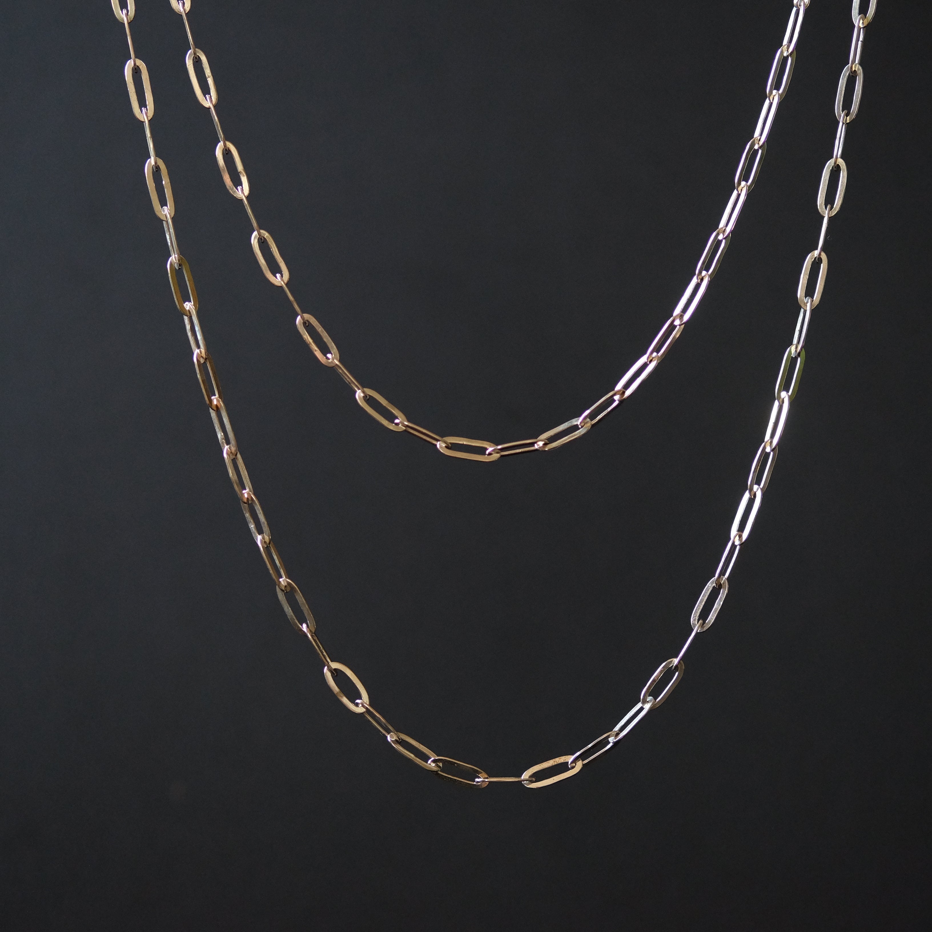 Two paperclip-link chain necklaces in gold and silver tones, hanging against a dark black background.