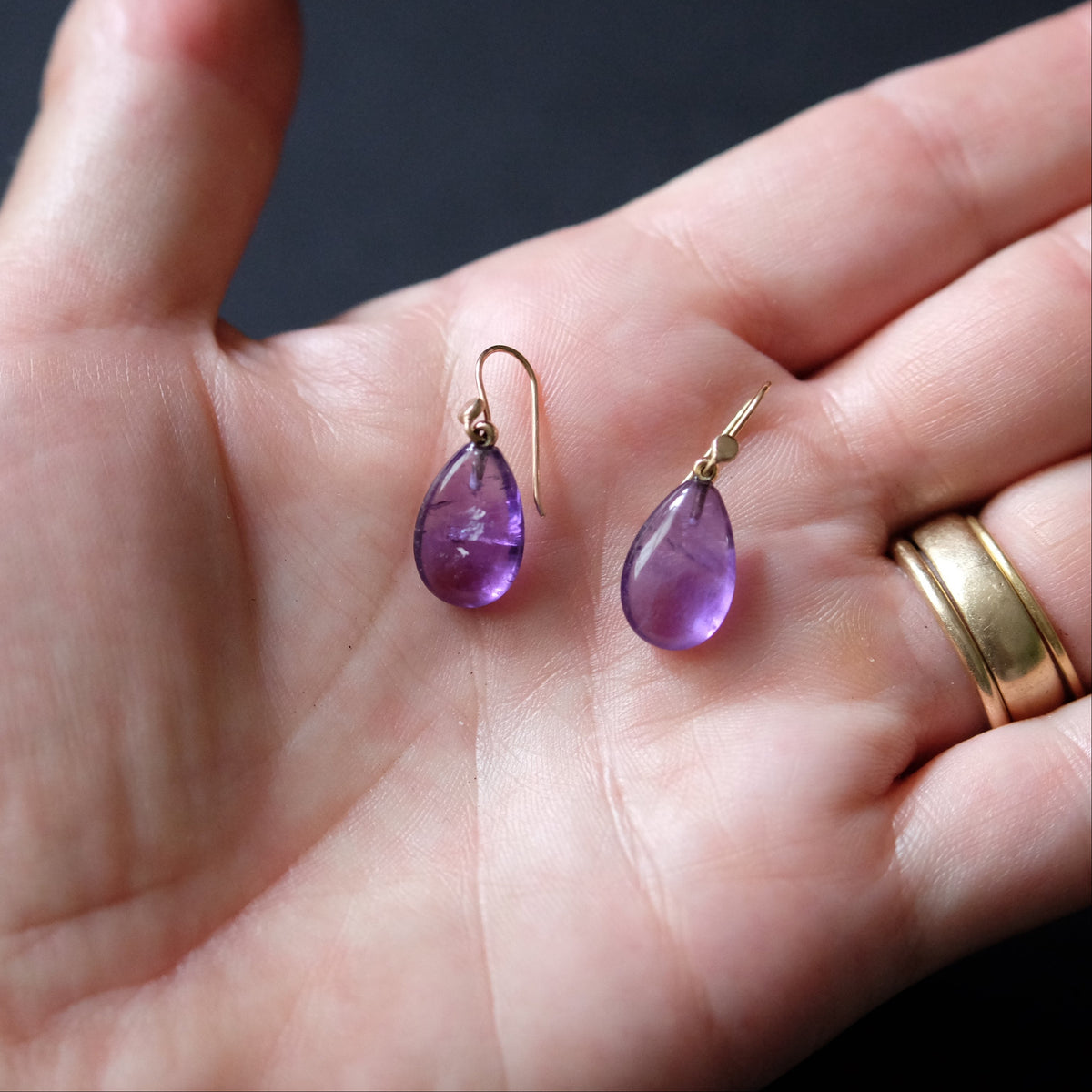 A pair of teardrop-shaped purple amethyst gemstone drop earrings with gold hooks, held in an open hand against a dark background.