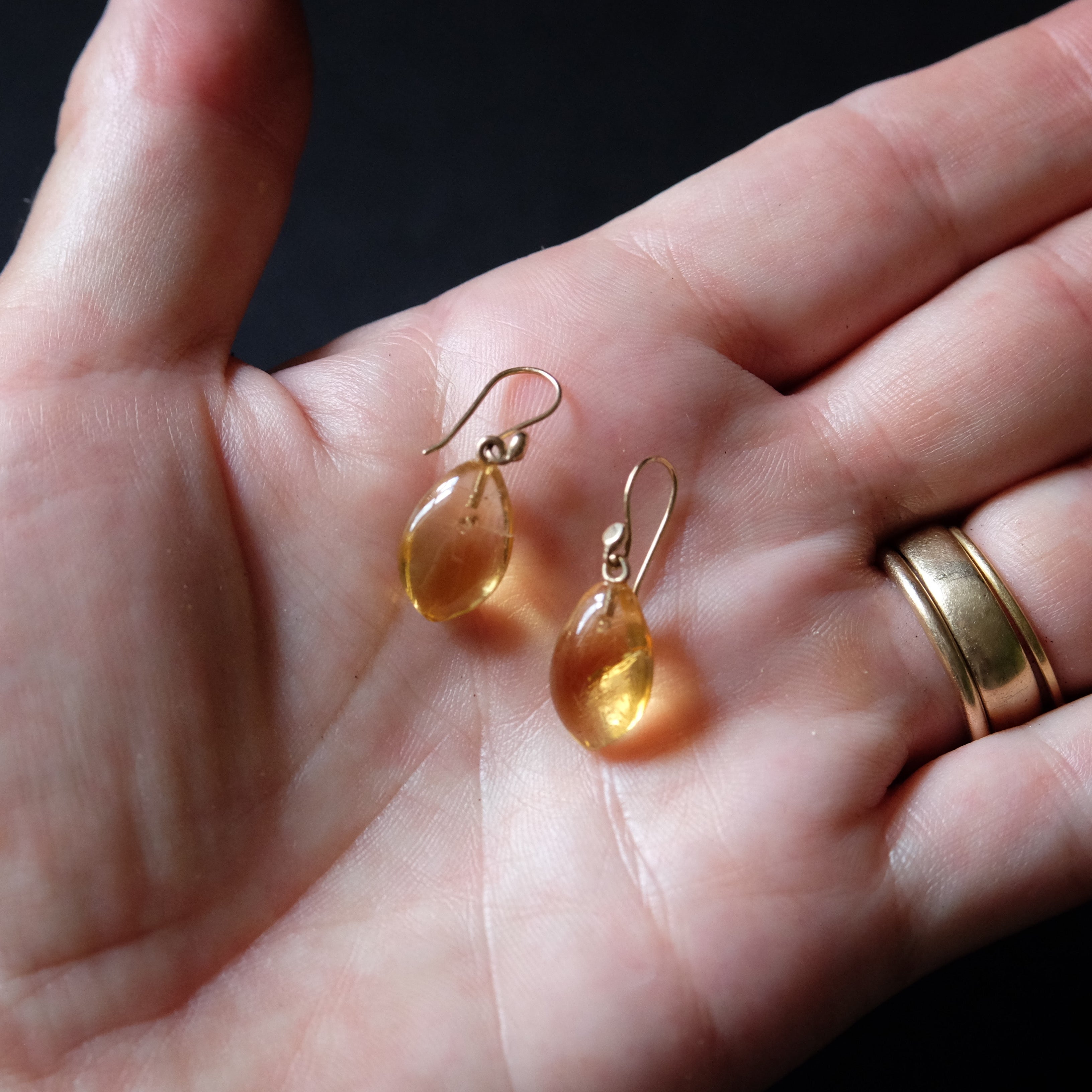 A pair of small teardrop-shaped amber glass drop earrings with gold fishhook wires, resting on an open hand next to a gold band ring.