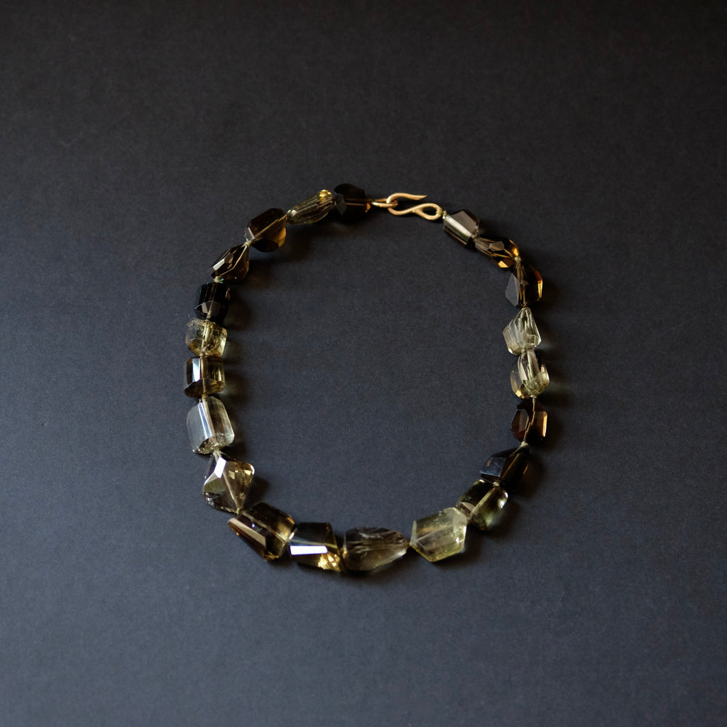 A beaded bracelet made of faceted translucent gemstone beads in smoky yellow, brown, and clear tones, with a gold-toned clasp, photographed on a dark gray background.