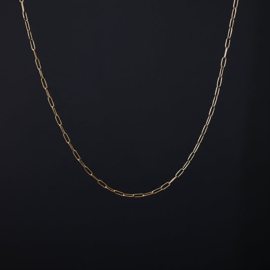 A delicate gold paperclip chain necklace draped in a U-shape against a dark textured background.