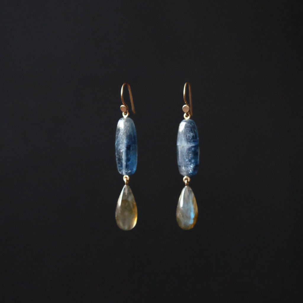 A pair of drop earrings featuring a polished blue stone bead and a teardrop-shaped labradorite stone, suspended from gold-toned hooks against a solid black background.