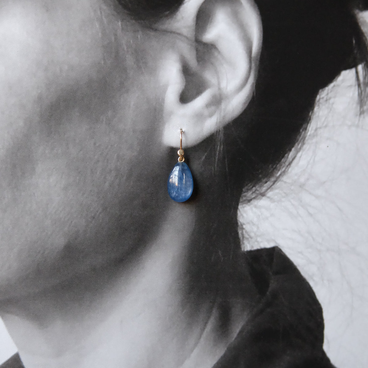 A close-up side profile of a person wearing a single teardrop-shaped blue drop earring with a gold fishhook back, shown in a black-and-white portrait with selective color on the earring.