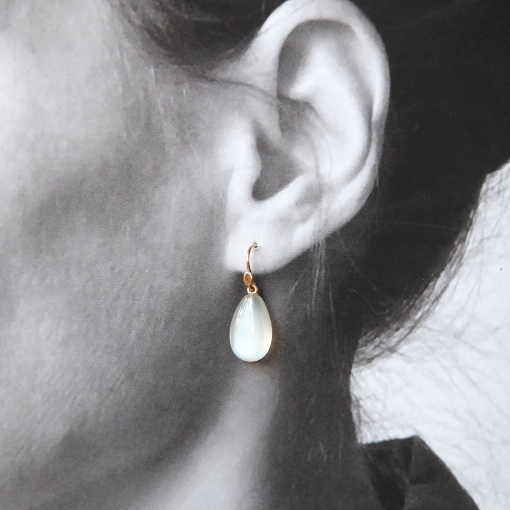 A close-up of a person's ear wearing a small gold hoop earring with a teardrop-shaped milky white stone drop.