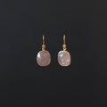 A pair of gold drop earrings with faceted light pinkish-purple stones, displayed against a dark black background.