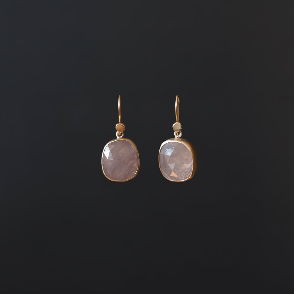 A pair of gold drop earrings with faceted light pinkish-purple stones, displayed against a dark black background.