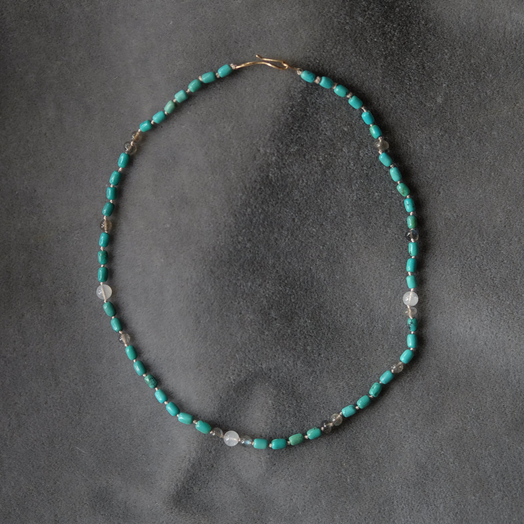 A delicate beaded necklace with turquoise blue beads, small silver spacer beads, and a few clear faceted beads, arranged in an oval on a dark gray textured background.
