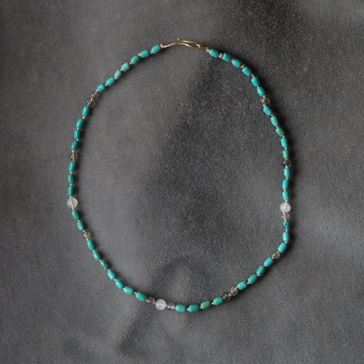 A delicate beaded necklace with turquoise blue beads, small silver spacer beads, and a few clear faceted beads, arranged in an oval on a dark gray textured background.