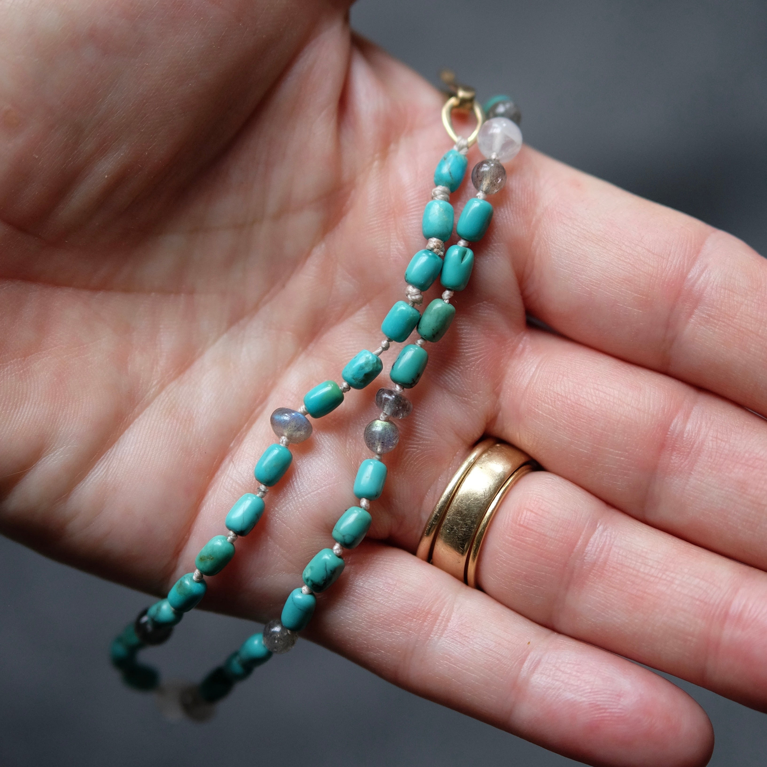 A close-up of a hand holding a beaded bracelet with turquoise-blue beads, small clear faceted beads, and gold-toned metal accents. The person is wearing a gold ring on one finger, and the background is a dark, blurred gray.