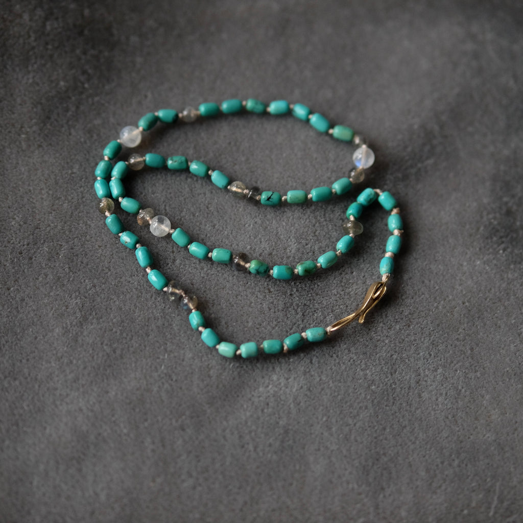 A beaded necklace featuring turquoise-colored beads, small gray stone beads, and a gold-toned clasp, displayed on a dark gray textured background.