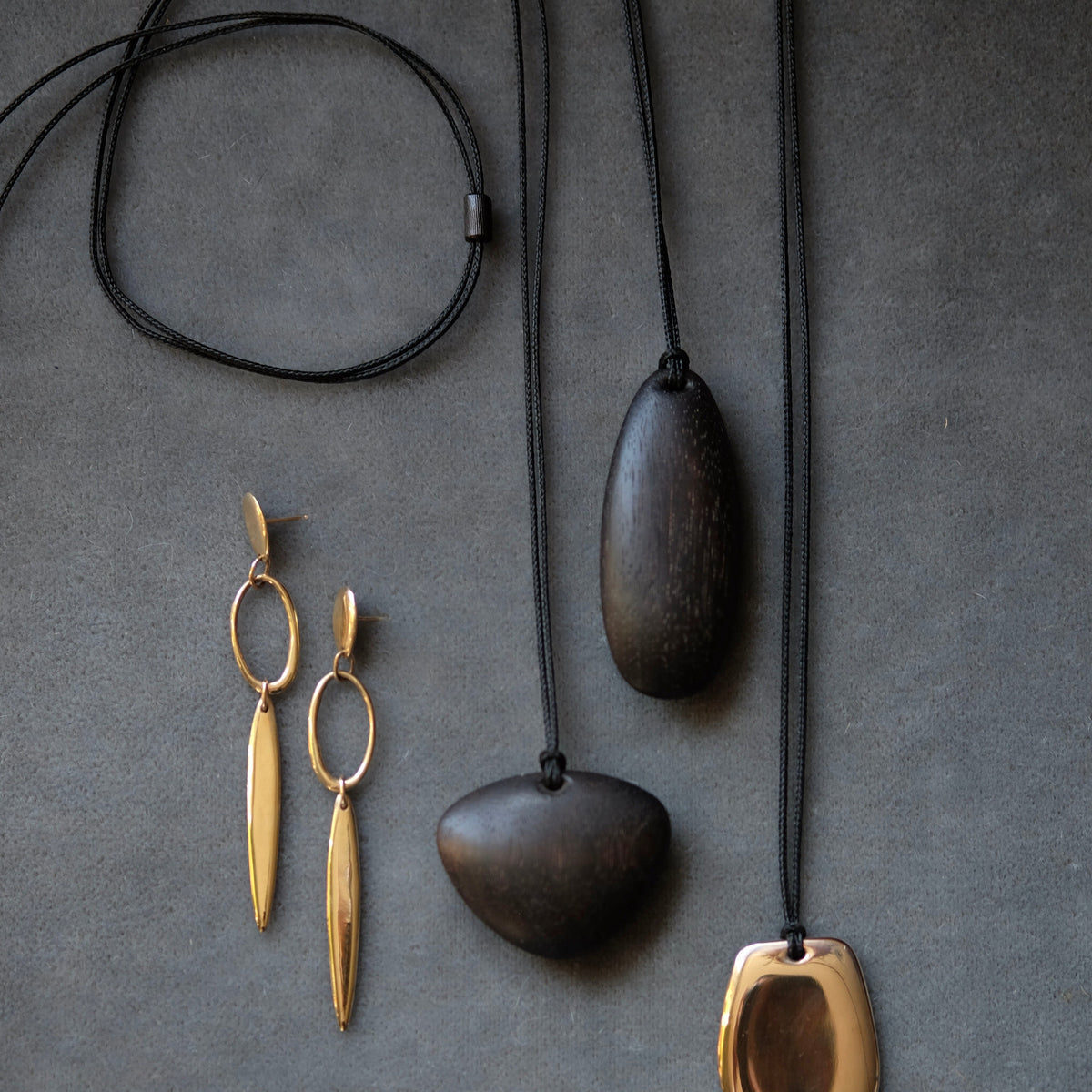 A styled flat-lay of minimalist jewelry on a dark gray textured background, featuring a black cord necklace with two dark wood pendants and a gold-toned pendant, alongside matching gold-toned drop earrings.
