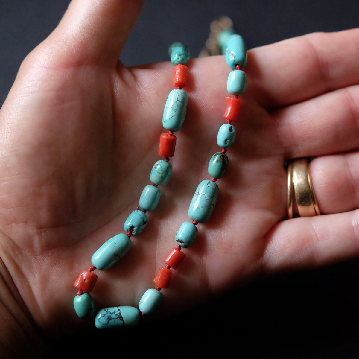 A beaded necklace with turquoise and coral-red beads draped over an open hand, with a gold ring visible on the finger against a dark background.