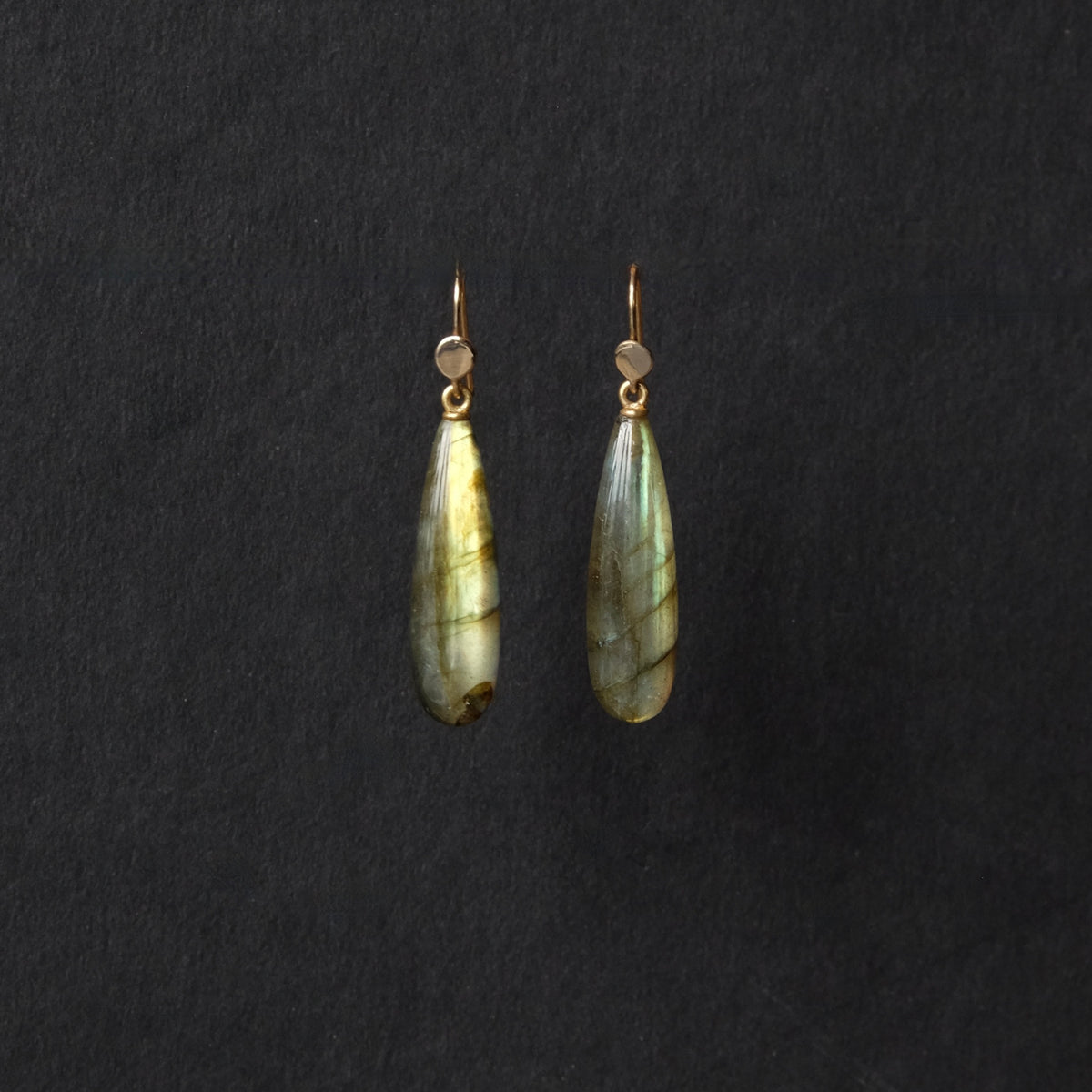 A pair of teardrop-shaped labradorite drop earrings with gold hooks, displayed against a dark textured background.