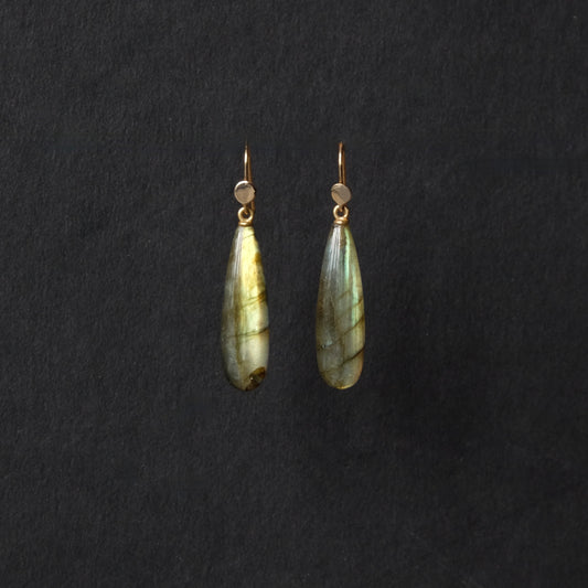 A pair of teardrop-shaped labradorite drop earrings with gold hooks, displayed against a dark textured background.