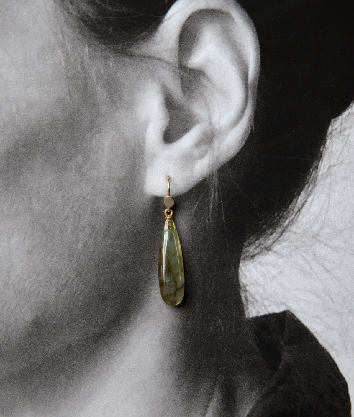 A close-up side profile of a person's ear wearing a single drop earring. The earring has a small gold-toned fishhook and bead with a teardrop-shaped olive green stone pendant.