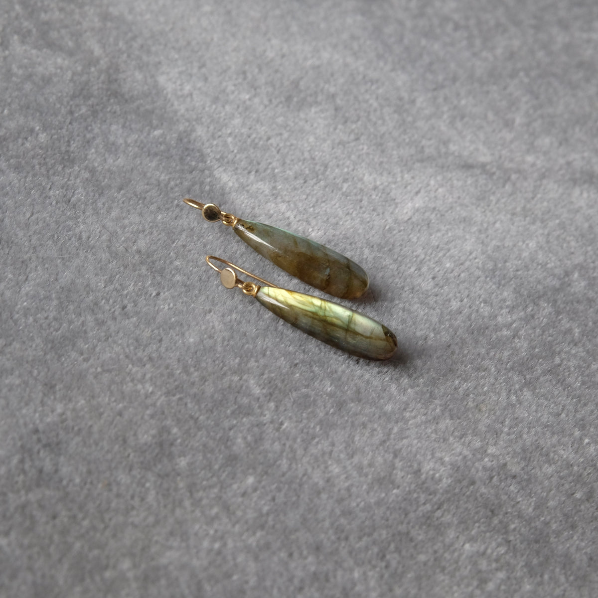 A pair of teardrop-shaped labradorite dangle earrings with gold-toned fishhook wires, resting on a textured gray fabric background.