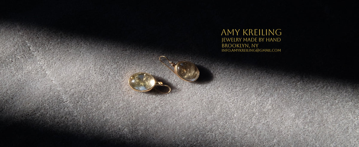 A pair of gold-toned drop earrings with oval clear stones, displayed on a textured gray surface under dramatic side lighting. The brand text 'AMY KREILING jewelry made by hand' appears in the upper right corner 