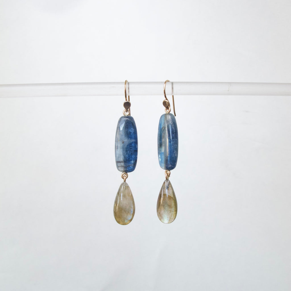 A pair of handmade dangle earrings featuring polished blue kyanite rectangular beads and teardrop-shaped smoky gray labradorite stones, suspended from gold-toned French hooks against a clean white background.