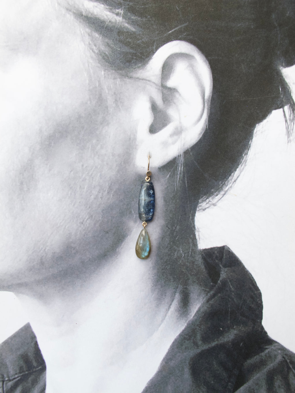 A close-up of a person's ear wearing a dangling earring with a dark blue stone and a smaller green teardrop charm, shown against a black-and-white portrait background.