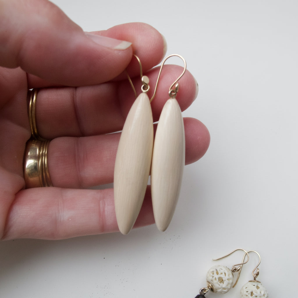 A hand holds a pair of light natural wood teardrop dangle earrings with gold fishhook wires. The background is a clean white surface with a second pair of dark wood and white bead dangle earrings lying in the lower right corner.