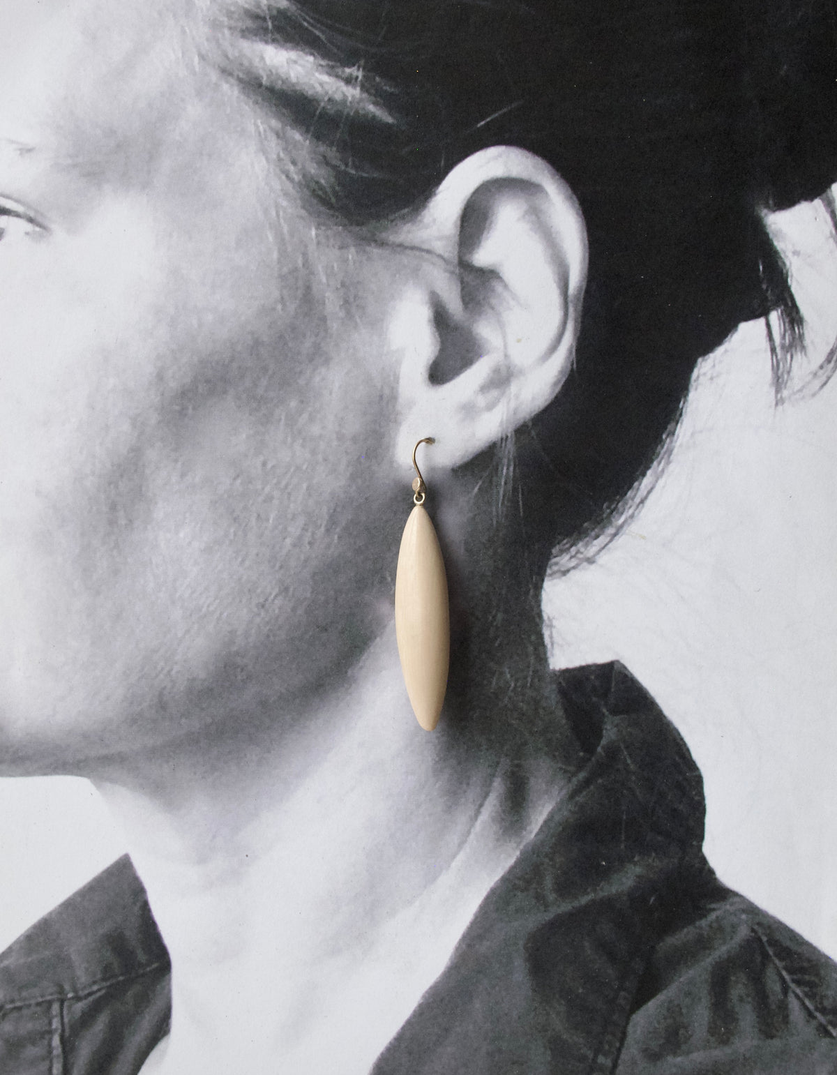 A close-up side profile of a person wearing a single teardrop-shaped cream-colored drop earring with a gold hook.