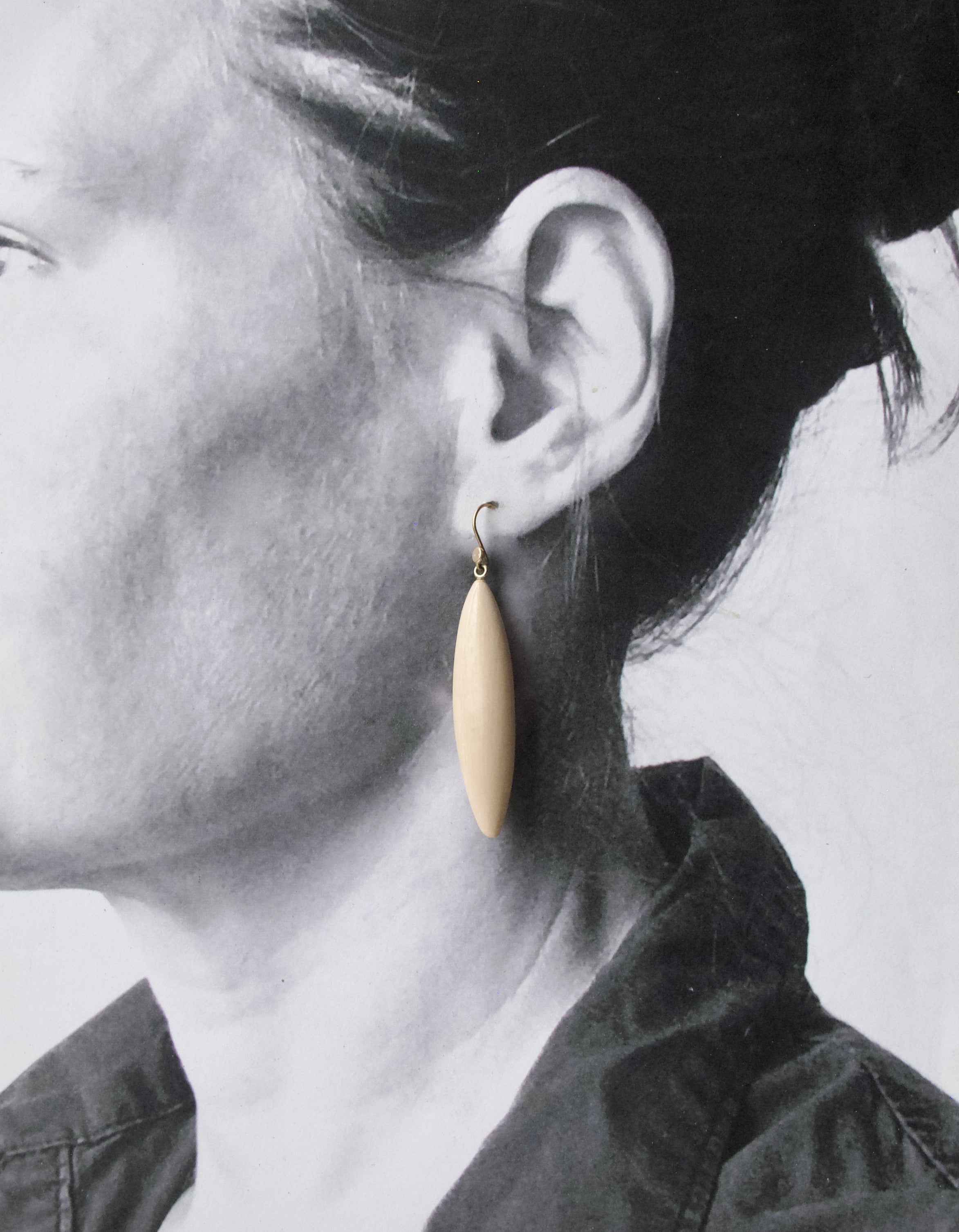 A close-up side profile of a person wearing a single teardrop-shaped cream-colored drop earring with a gold hook.