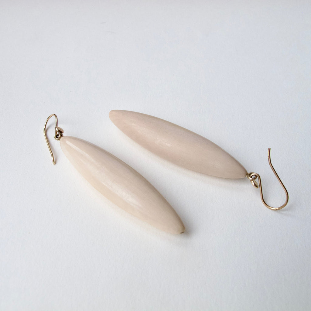 A pair of minimalist dangle earrings featuring smooth, elongated oval beads in a light cream-beige color, suspended from simple gold-tone French hook wires, set against a clean white background.