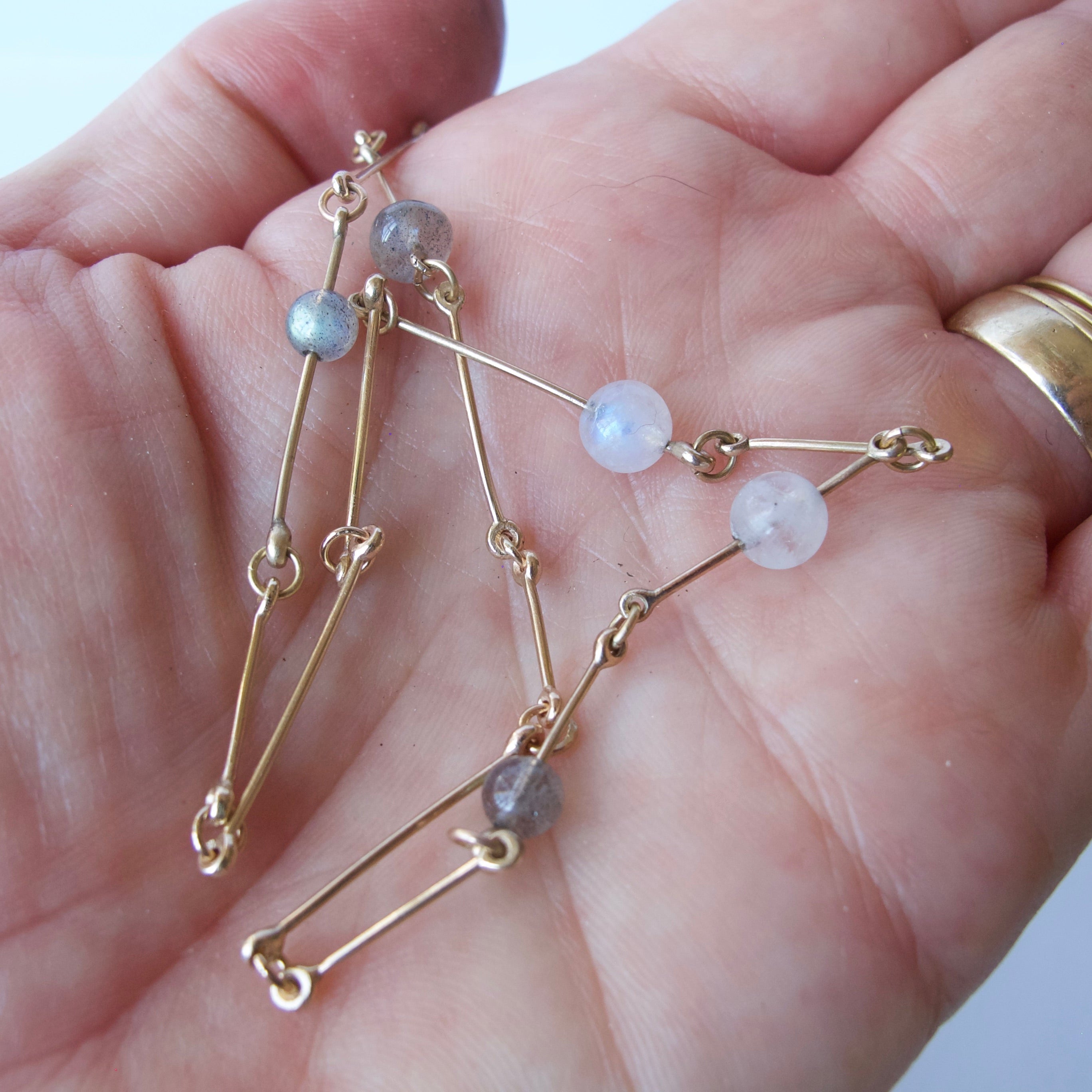 A delicate gold chain necklace with small gray and pale blue beads, held in a person's hand. The person is wearing a gold band on their right hand.