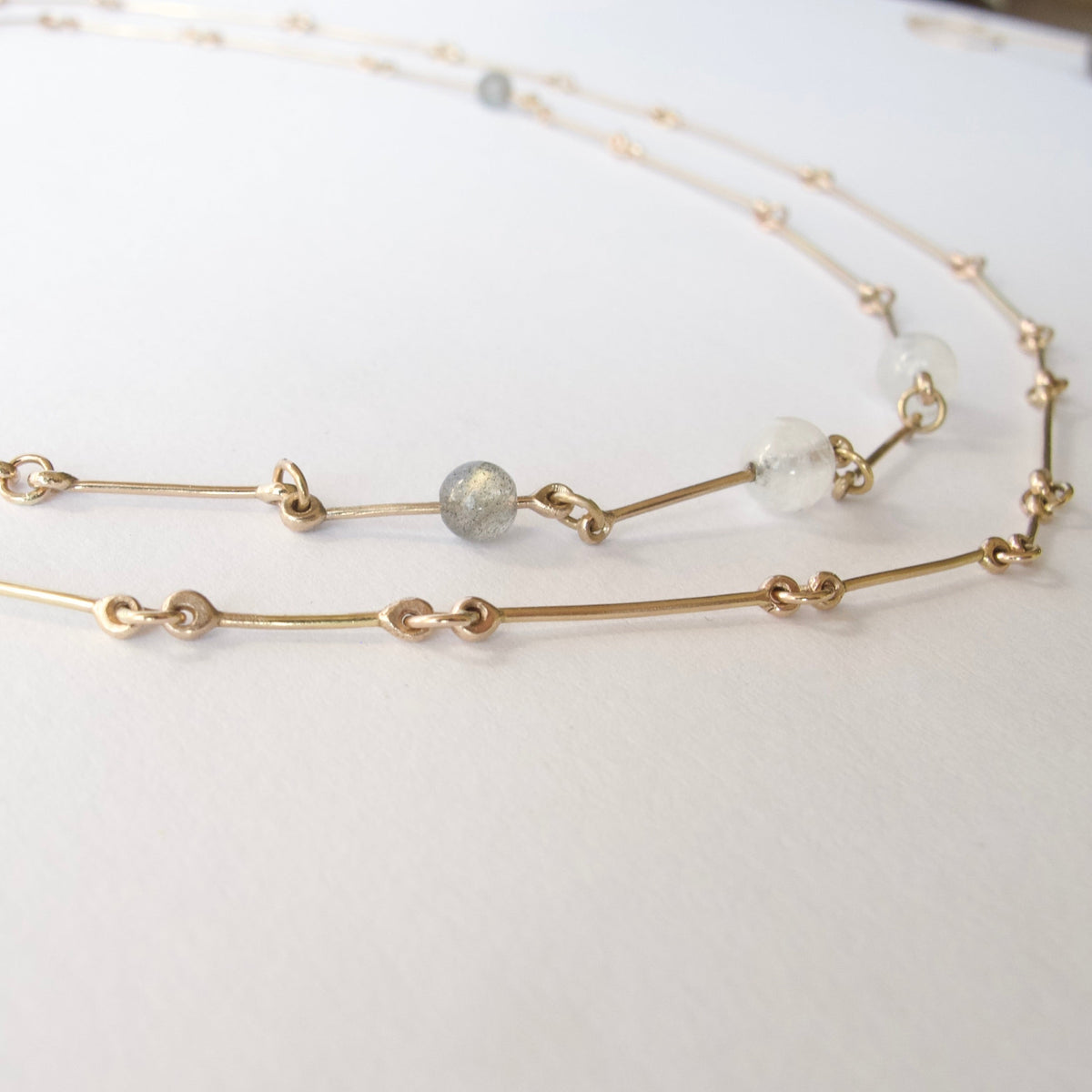 A delicate gold chain necklace with small bead links and two clear faceted beads, displayed on a plain white surface.