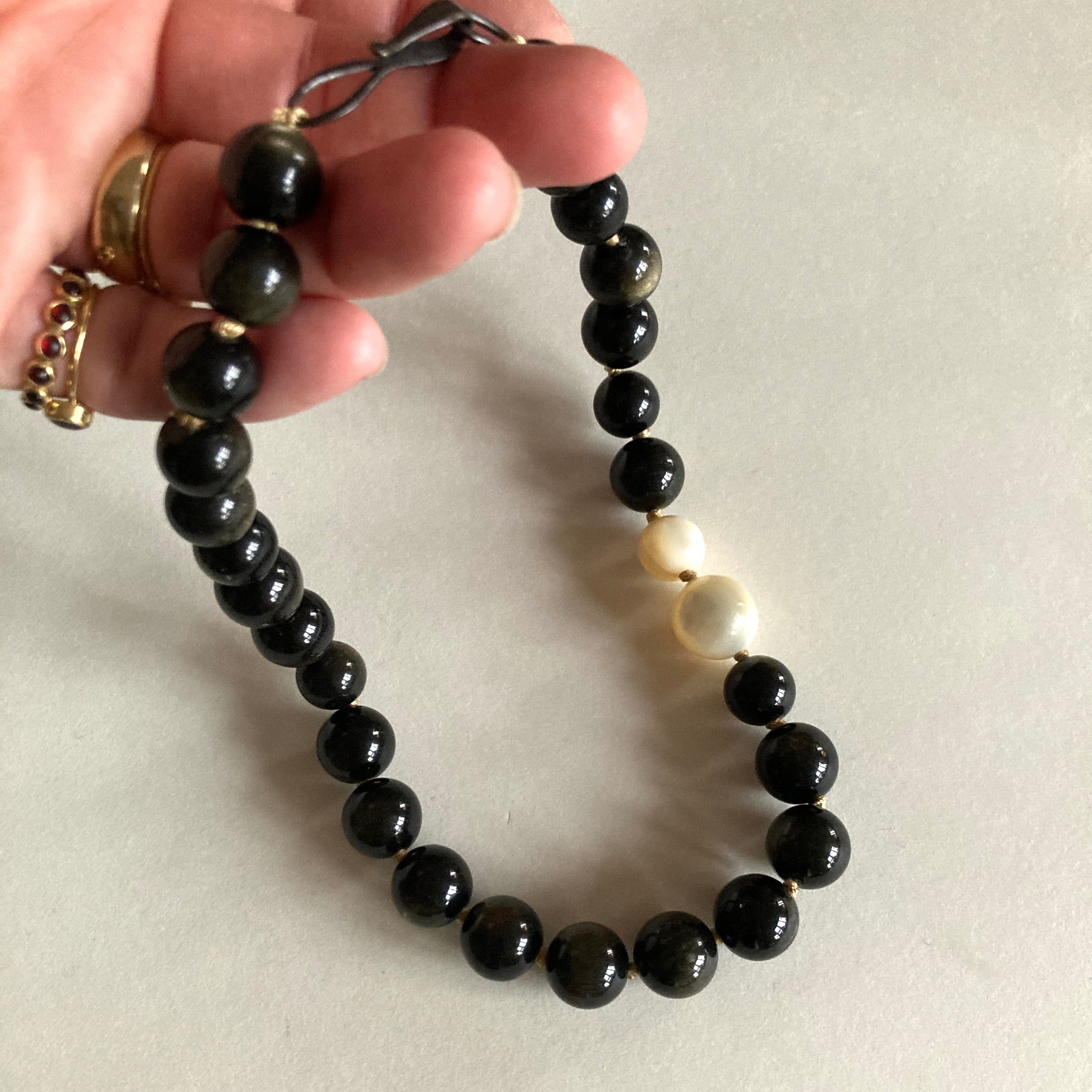 A beaded necklace with glossy black round beads, gold spacer accents, and one prominent white bead, held by a hand against a plain off-white background.