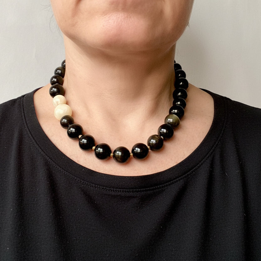 A close-up of a person wearing a beaded necklace with mostly dark polished beads and one light cream bead, paired with an olive green top against a plain light background.