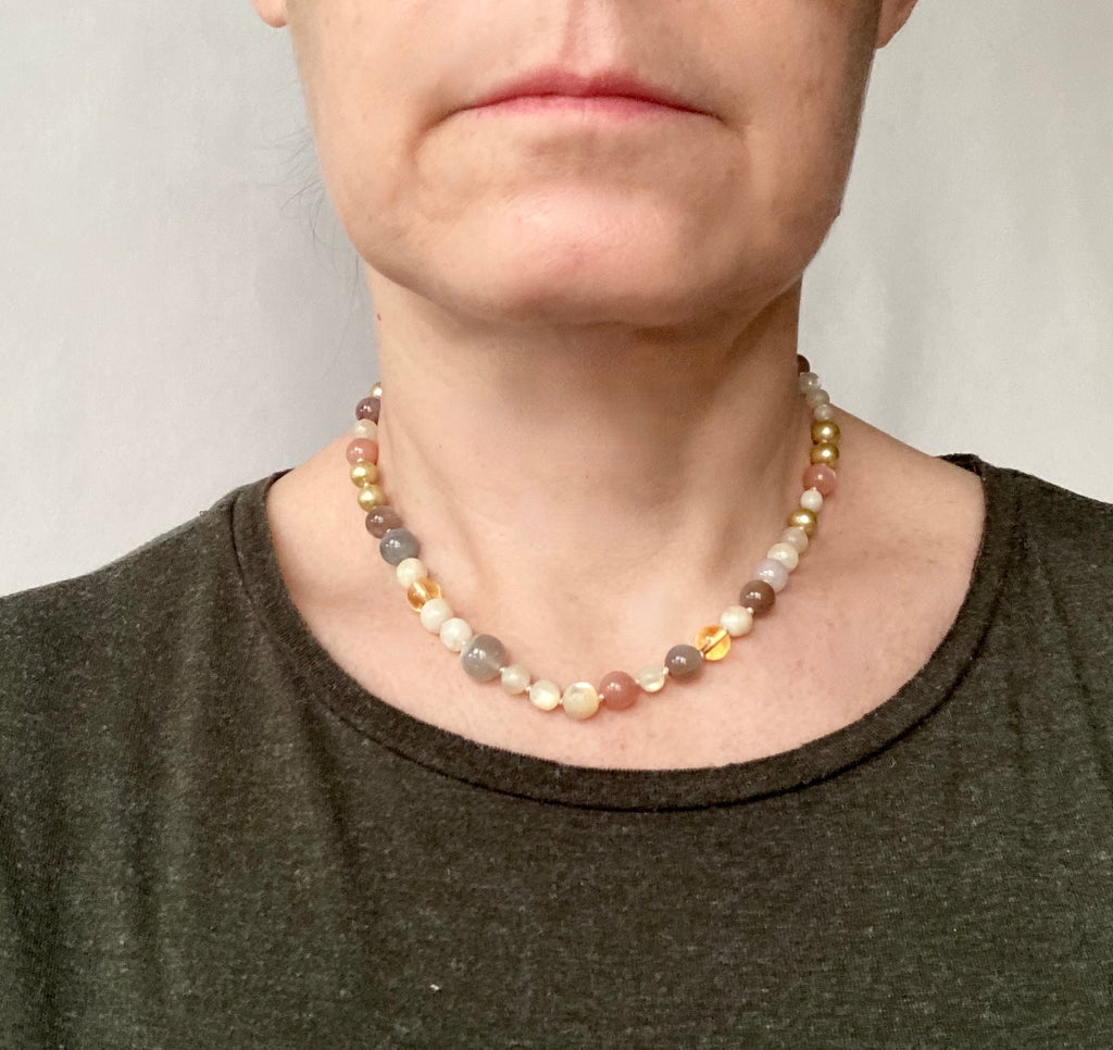 A close-up of a person wearing a multi-colored beaded necklace with gray, cream, orange, and gold-toned beads, paired with a dark olive green shirt.